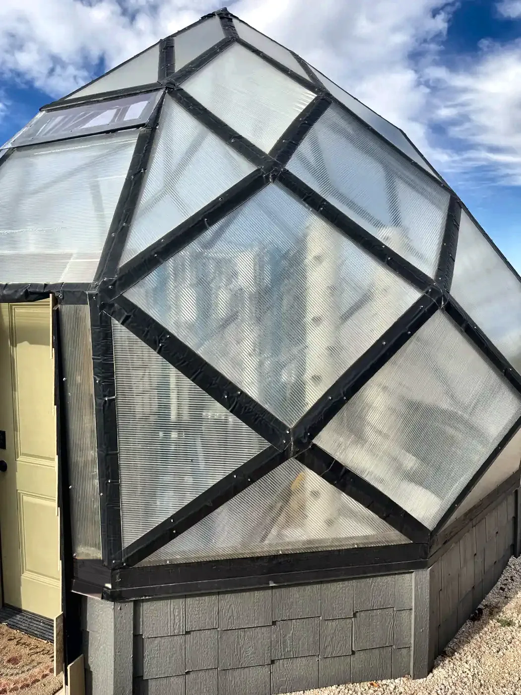 The zome greenhouse from outside: frosted polycarbonate panels in black frames forming a spiral of diamonds above a gray shingled pony wall.