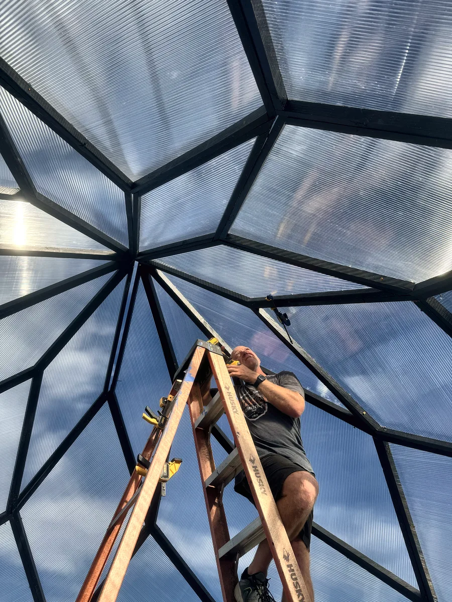 Looking up from inside during the build: a person on a ladder working at the apex as the panels close in.