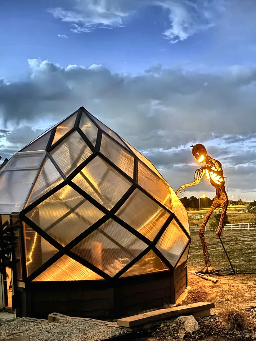 The zome glowing warm amber at dusk beside a metal sculpture and a tree.