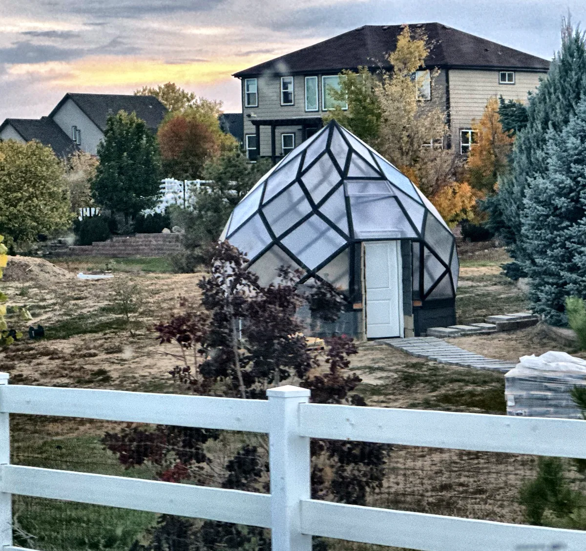 The zome in a Brighton backyard behind a white rail fence, with houses and evergreens around it.